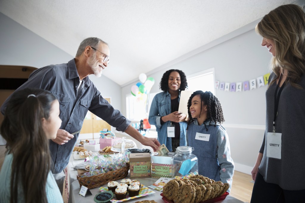 Adults and kids surrounding a table of baked goods. Sign taped on wall that says "BAKE SALE"