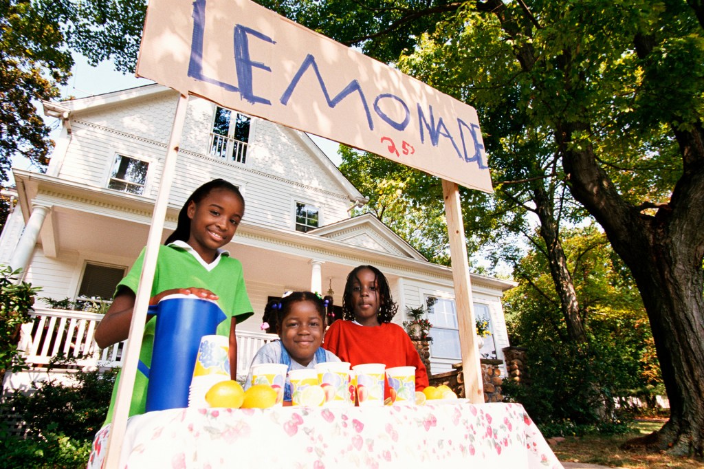 three kids smile from behind a lemonade stand