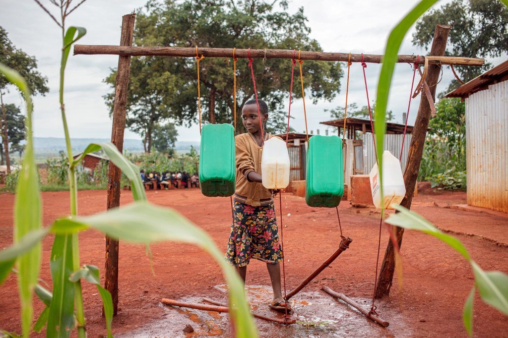 kid standing behind four green and white jugs of water dangling on wood stick by strings