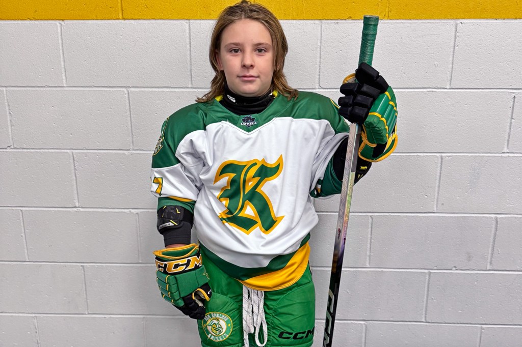 A young hockey player in a green and white uniform stands against a wall, holding a hockey stick and looking at the camera.