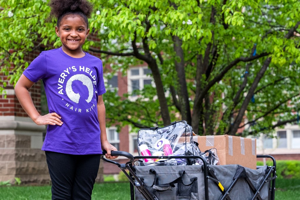 A smiling child wearing a purple T-shirt stands outdoors beside a wagon filled with supplies.