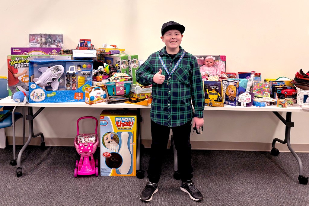 A smiling child stands in front of a table covered with toys, giving the thumbs up.