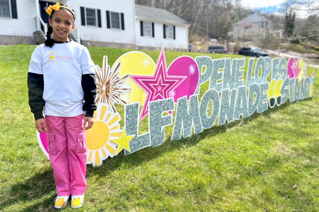 A smiling child stands outdoors in front of a colorful sign that reads “Penelope’s Lemonade Stand.”