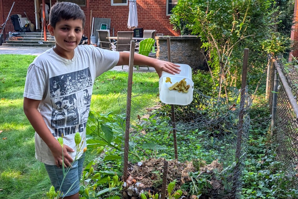 A child stands in a backyard garden, holding a container of food scraps over a compost pile.