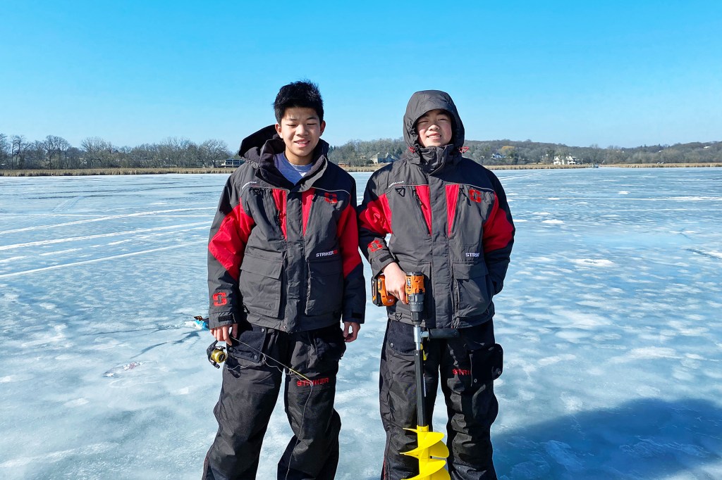 Two kids wearing winter gear stand on a frozen lake. One holds a fishing rod, the other drill with an ice auger.