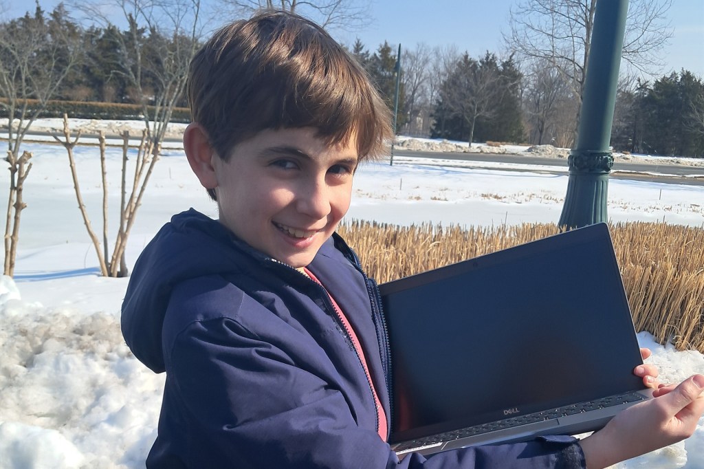 A smiling boy in a winter jacket stands outside in the snow, holding a laptop open toward the camera.