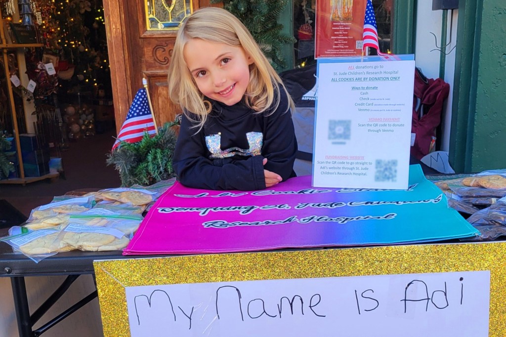 A smiling girl sits at a table selling cookies to raise money for a children's hospital.