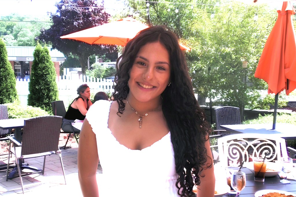 A smiling young woman with long, curly hair standing in an outdoor dining area.