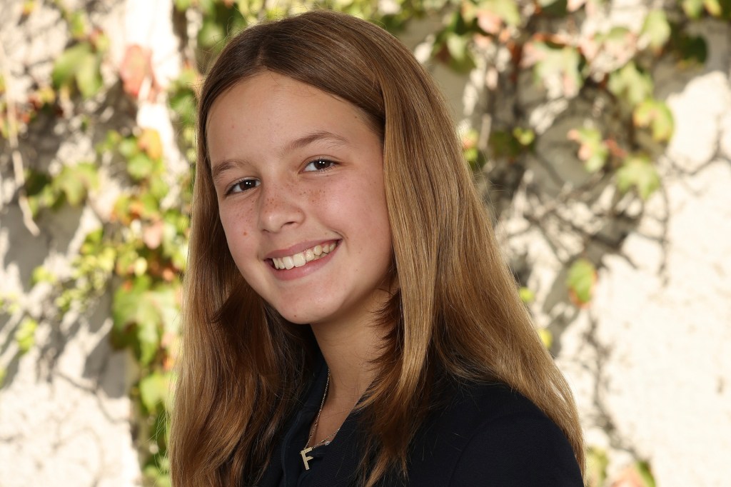 A young person with long light brown hair smiles at the camera while standing outdoors in front of a wall covered with green and brown ivy.