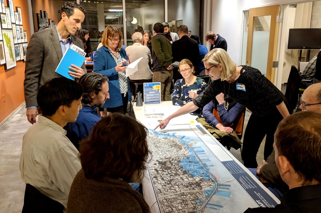 A group of people around a table looking at a map of a city. A woman on the right points to a spot on the map.
