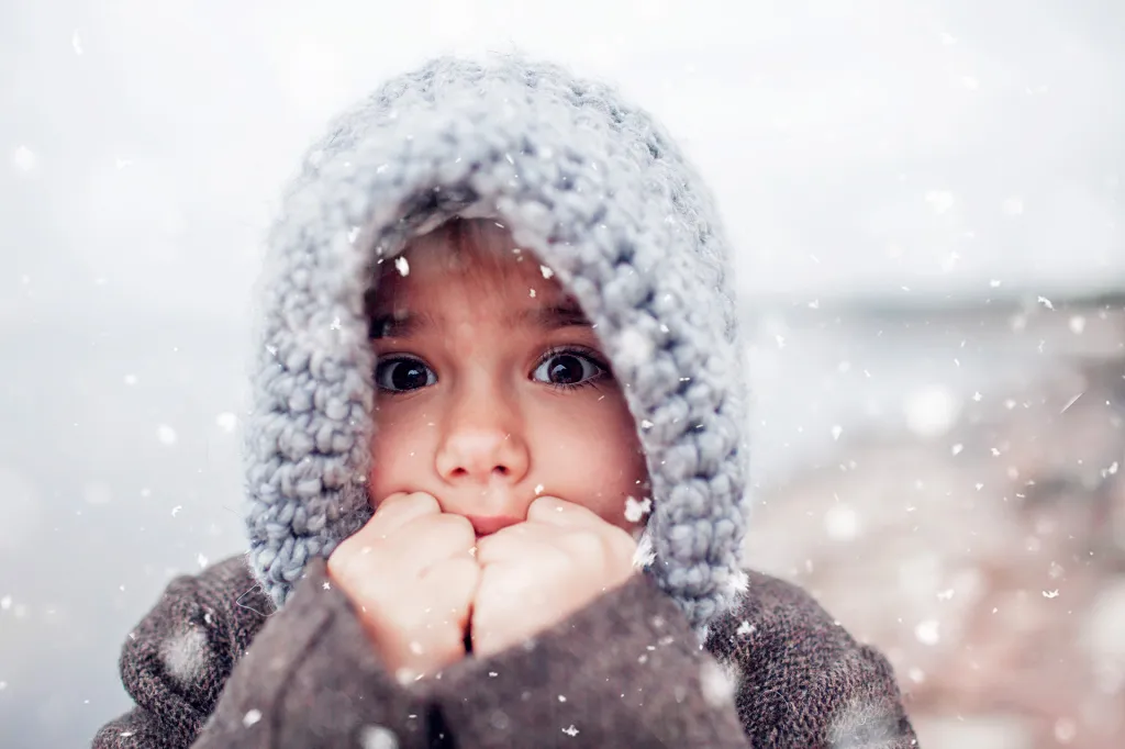 Close-up of a child in a gray knit hood and brown coat standing in falling snow, holding their hands near their face.