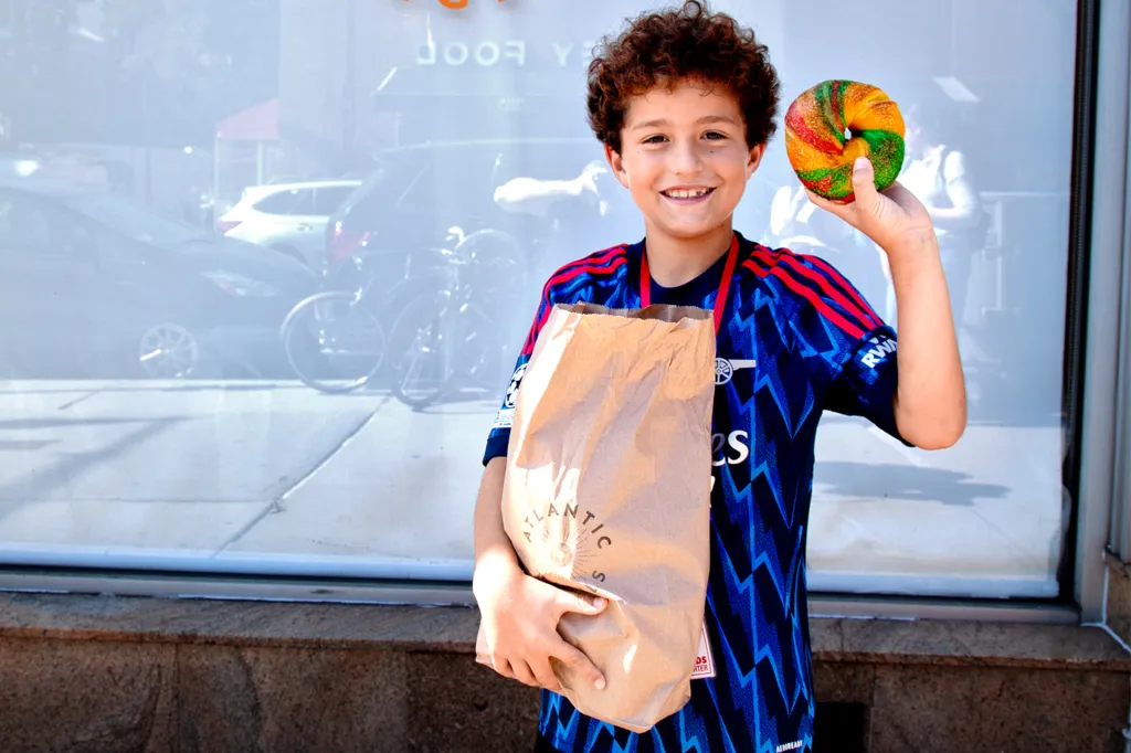 Smiling boy in a blue soccer jersey holds up a colorful bagel and a brown paper bag outside a shop window.
