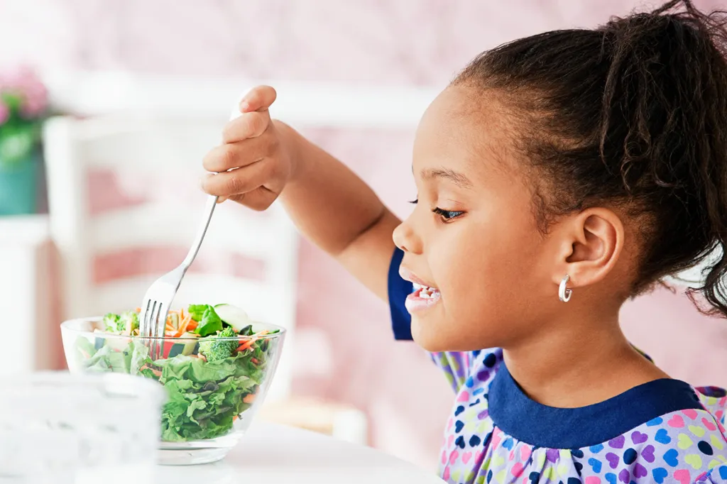 Girl smiles as she eats a fresh salad from a glass bowl at a table.