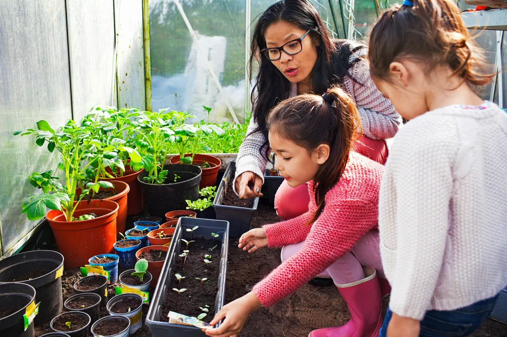Adult and two children plant seedlings in soil trays inside a greenhouse filled with potted plants.