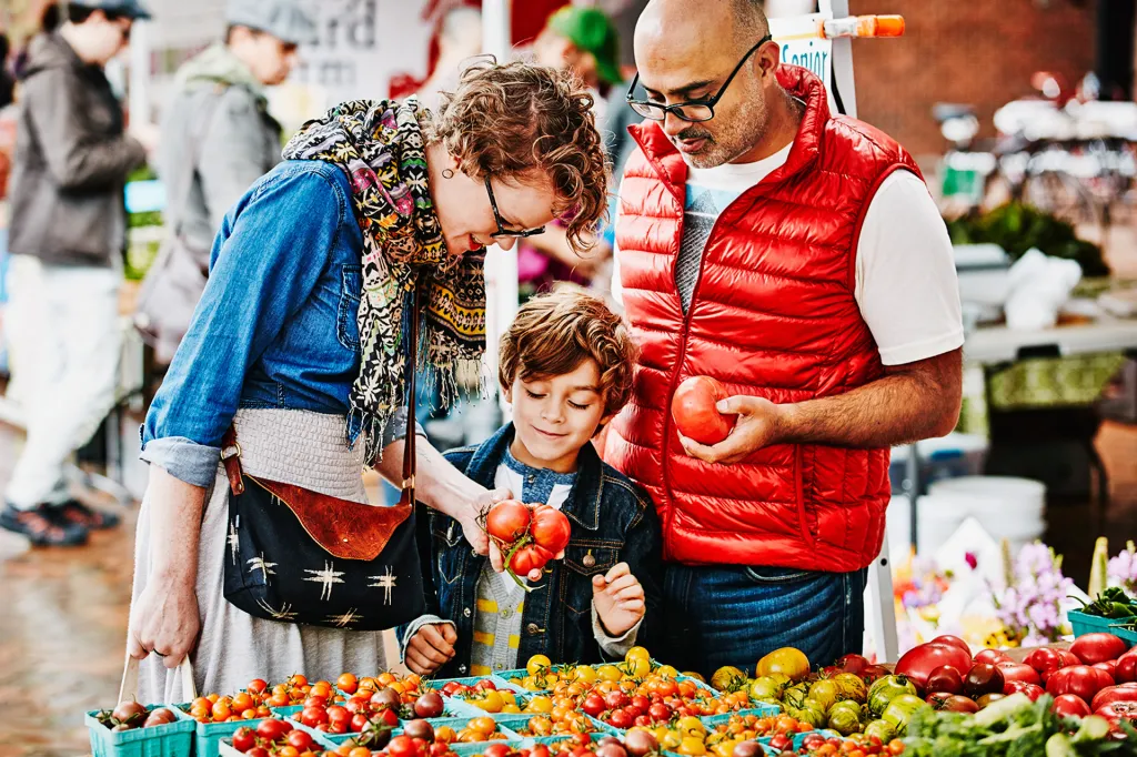 Family shops for fresh tomatoes at a farmers market, standing in front of colorful produce displays.