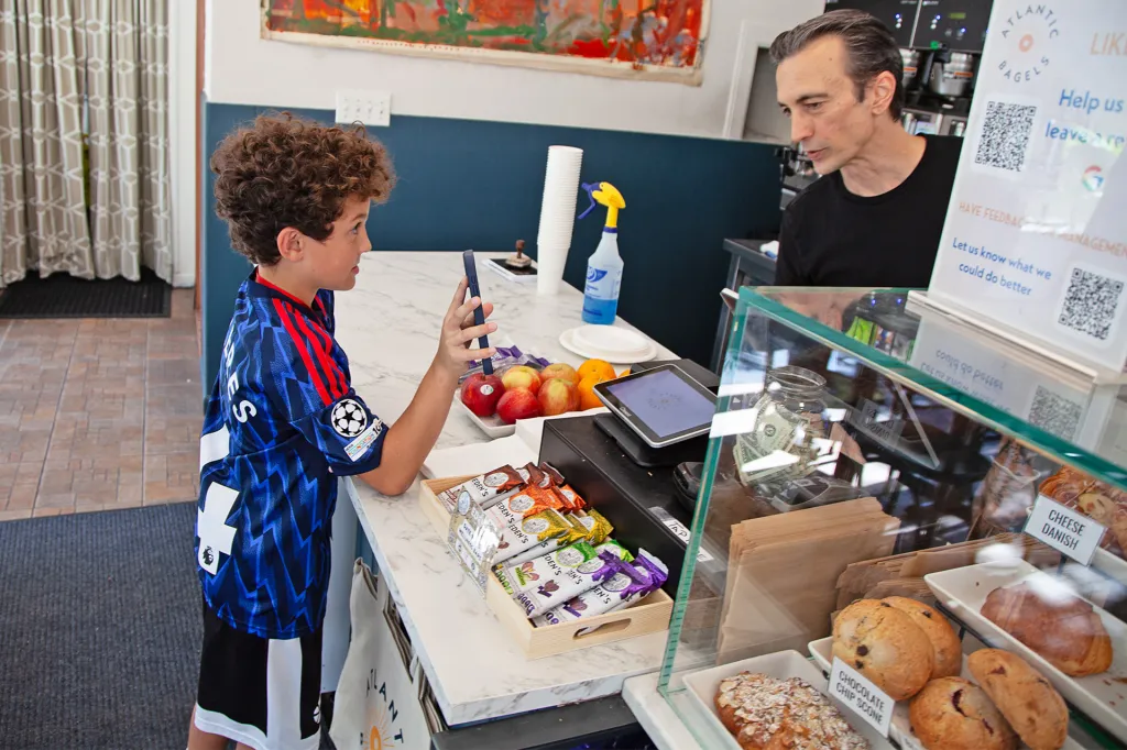 Boy in a blue soccer jersey uses his phone to order food at a counter inside a bagel shop.