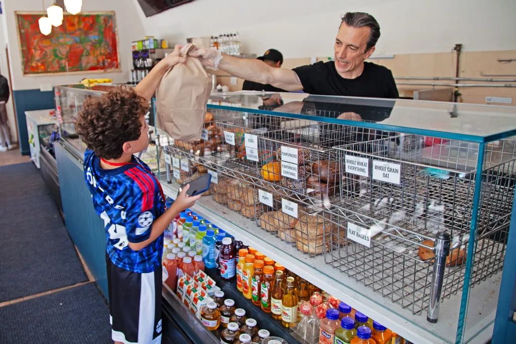 Shop employee hands a paper bag of bagels to a boy in a blue soccer jersey standing at the counter.