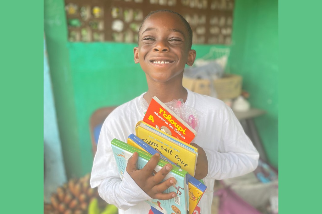 Smiling boy holds a stack of colorful children’s books in front of a green wall.