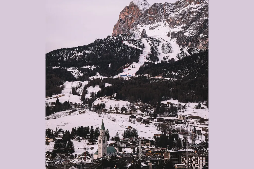 Snow-covered mountains rise above the town of Cortina d’Ampezzo, Italy, with a church steeple in the foreground and ski slopes winding down the hills.