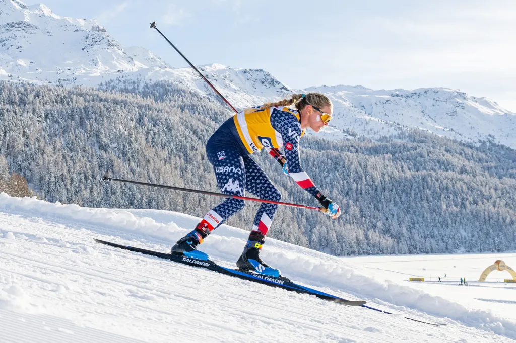 A cross-country skier wearing a blue and yellow uniform races across a snowy slope with forested mountains in the background.