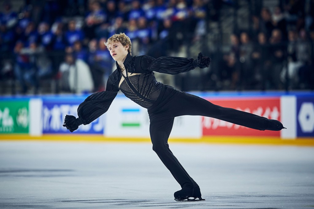 A figure skater glides across the ice with one leg extended behind him and arms outstretched, performing in front of an audience.