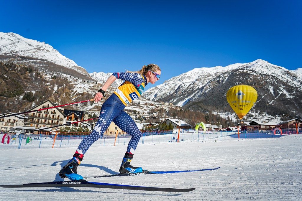 A cross-country skier races through a snowy course surrounded by mountains, wearing a blue and yellow suit and using ski poles.