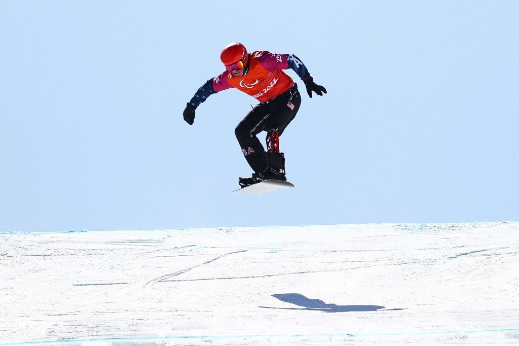 A snowboarder wearing a red helmet and Paralympic gear soars through the air above a snowy slope.