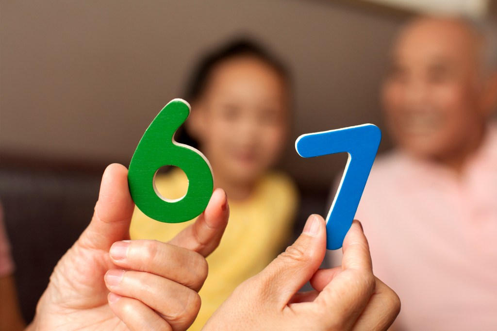 Two hands hold up colorful wooden numbers 6 and 7, with a child and an older adult smiling in the blurred background.