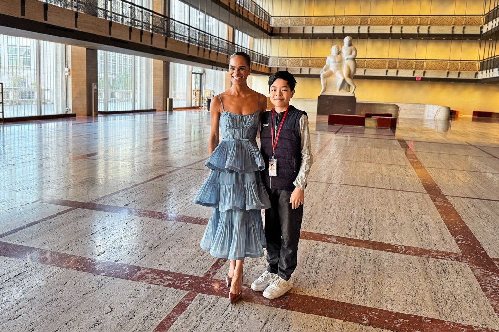 Smiling woman in a light blue layered dress stands beside a boy wearing a black vest inside a large, sunlit building.