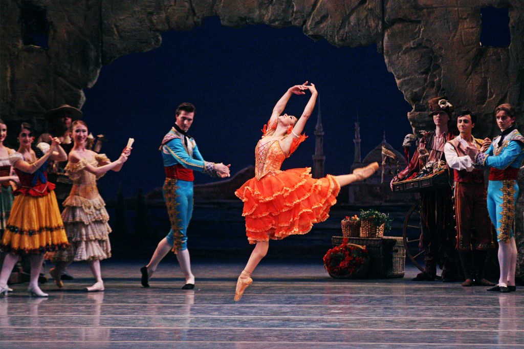 Ballerina leaps across the stage in an orange dress during a performance, with other dancers watching in the background.