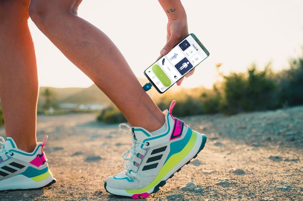 A person on a rocky trail holds a smartphone connected to a small device on their ankle, with a fitness app open on the screen.