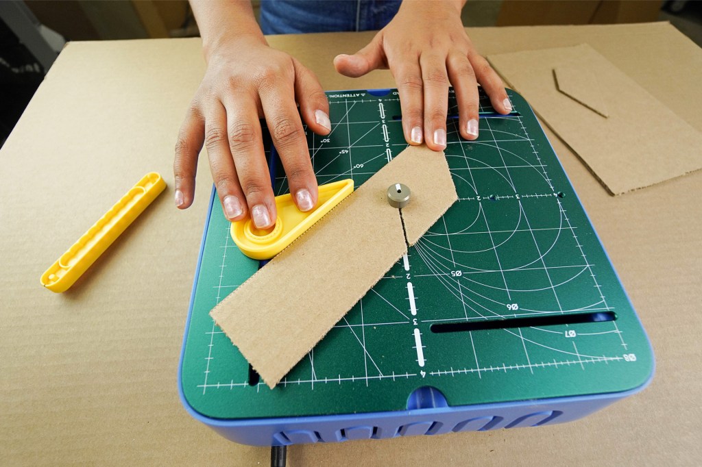 Hands use a small cutting tool to shape a piece of cardboard on a green crafting board.