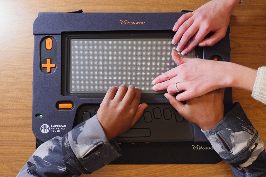 Two pairs of hands touch the raised dots on a braille display labeled “Monarch.”