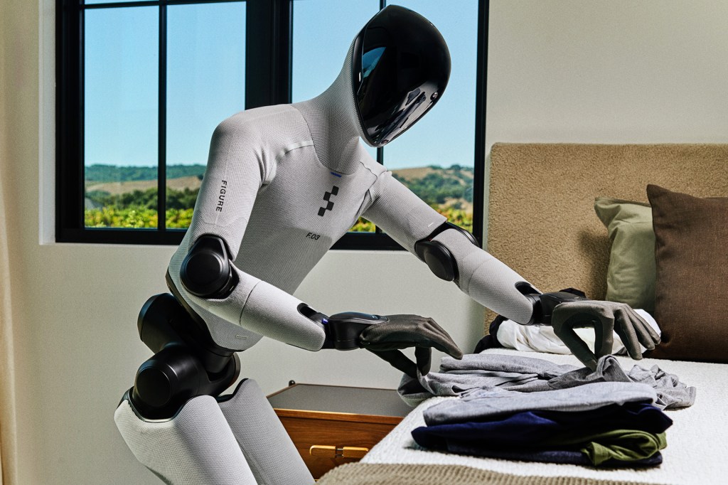 A humanoid robot folds clothes on a bed inside a modern, sunlit room.