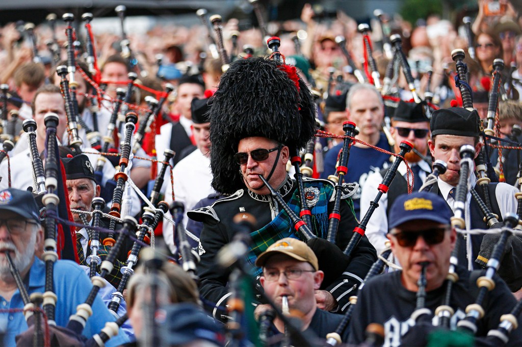 A large group of people play bagpipes in a parade, with one man wearing a tall black feathered hat leading in the center.