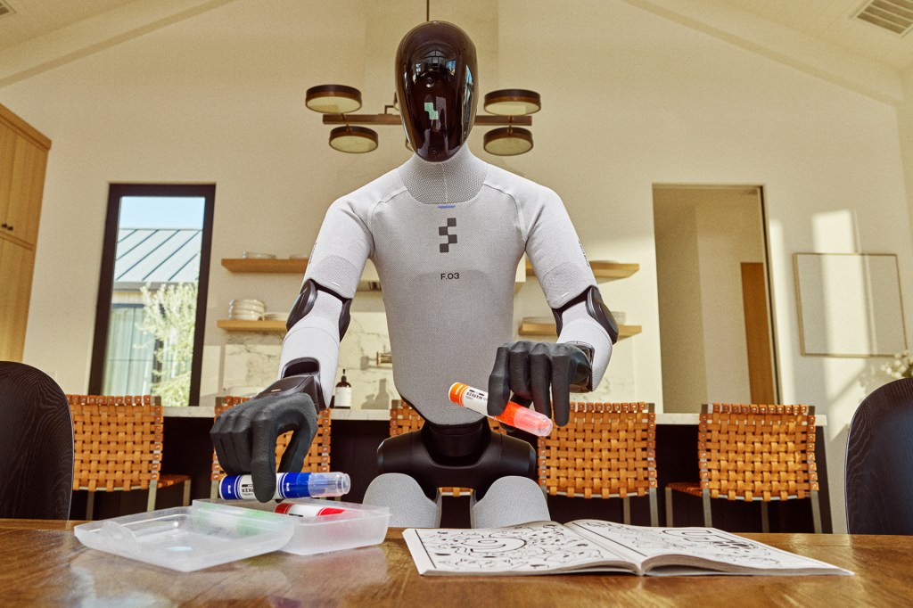 A humanoid robot stands near a table in a modern kitchen, holding colored markers above an open coloring book.