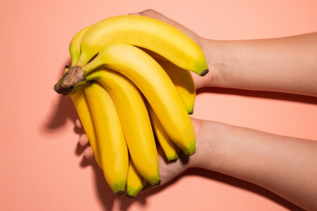 Two hands hold a bunch of bright yellow bananas against a peach-colored background.
