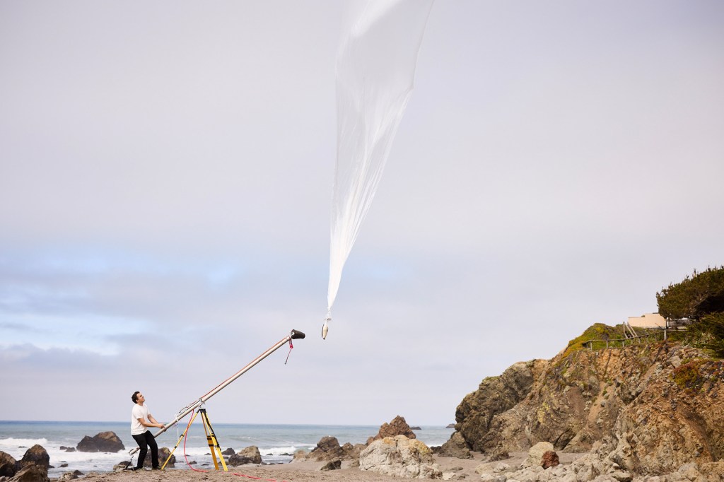 A person stands on a rocky beach launching a large white weather balloon into the sky.