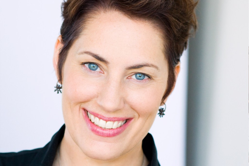 A woman with short brown hair smiles at the camera. She is wearing small black earrings and a black collared shirt against a light background.