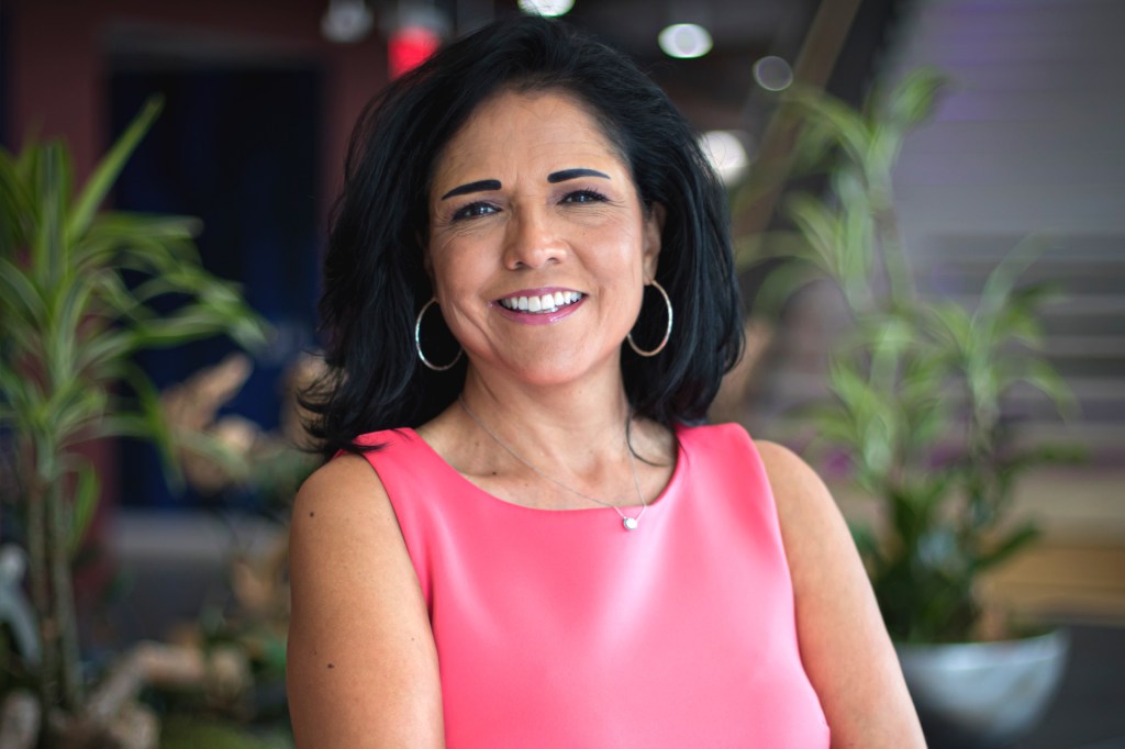 A smiling woman with dark hair wears a bright pink sleeveless top and hoop earrings, standing indoors with plants in the background.