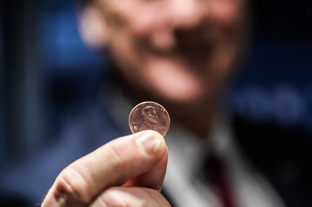 Hand holding a U.S. penny; person blurry in background.