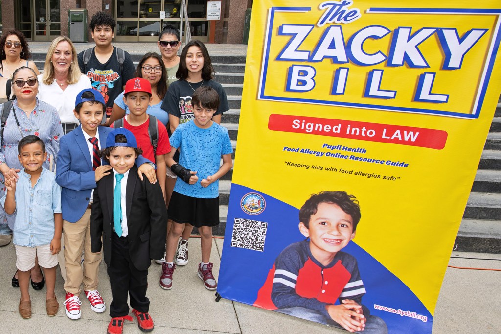 A group of kids and adults stands by a yellow banner: “The Zacky Bill—Signed into LAW.”