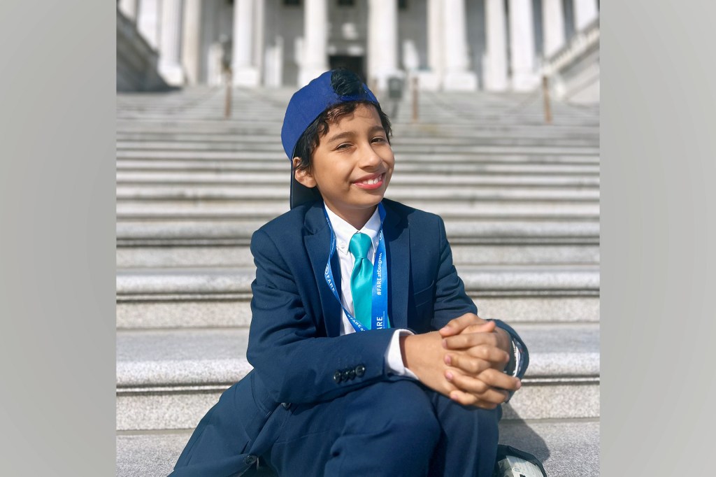 Kid in suit and blue cap sits smiling on Capitol steps.