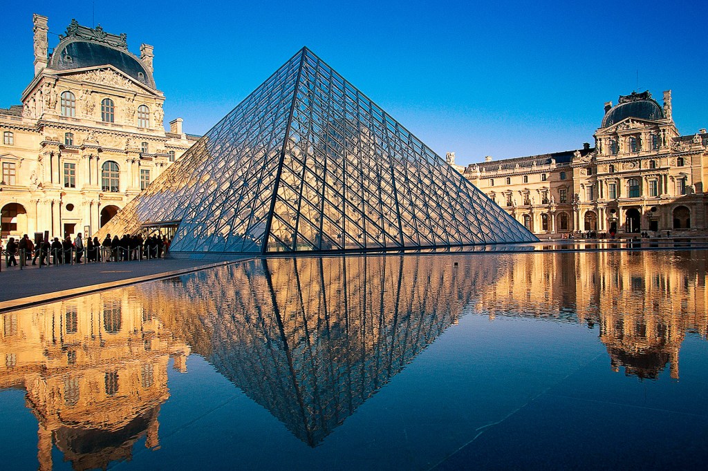 The Louvre’s glass pyramid reflects in a pool beside historic buildings.