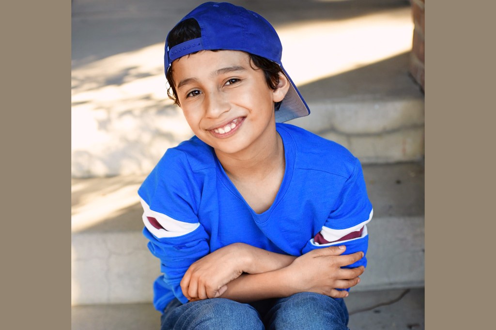 A smiling kid in blue shirt and cap sits on steps.