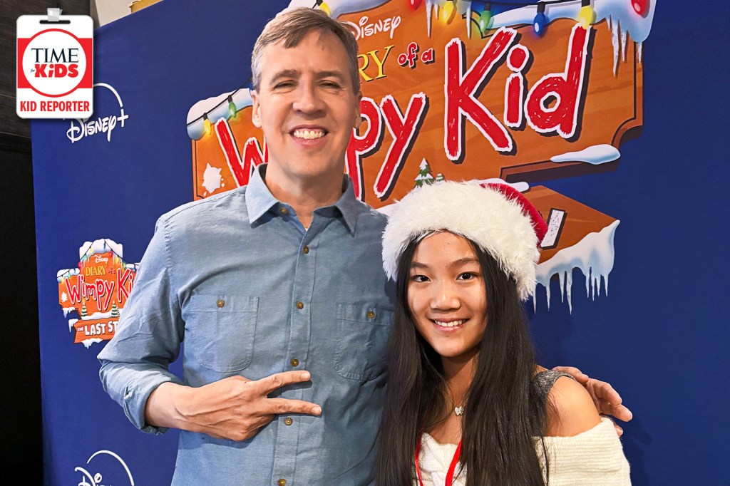 A smiling man stands next to a smiling girl wearing a Santa hat. They pose in front of a bright “Diary of a Wimpy Kid” movie poster; the man makes a peace sign and the girl looks at the camera.