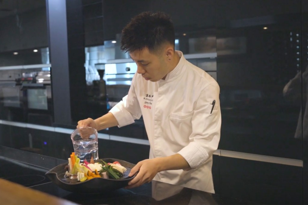 A chef in a restaurant kitchen pouring a liquid into a bowl of colorful food.