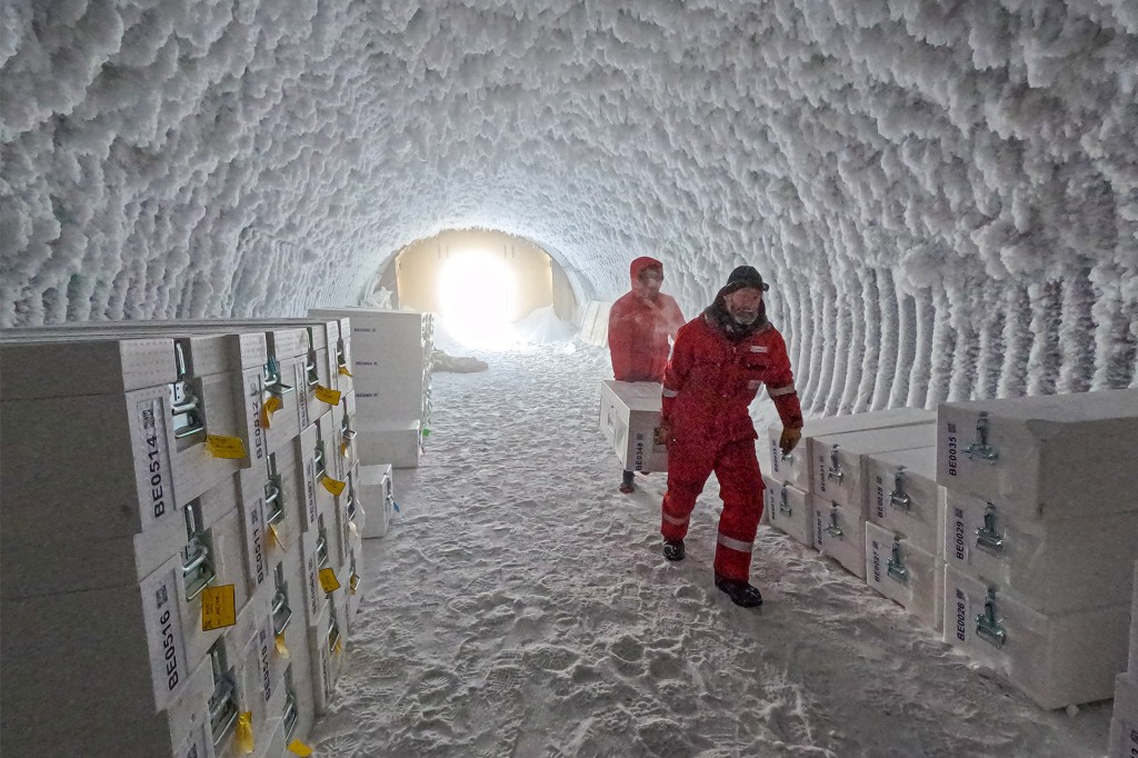 Two people in red cold-weather suits carry white boxes through a tunnel carved out of snow and ice.
