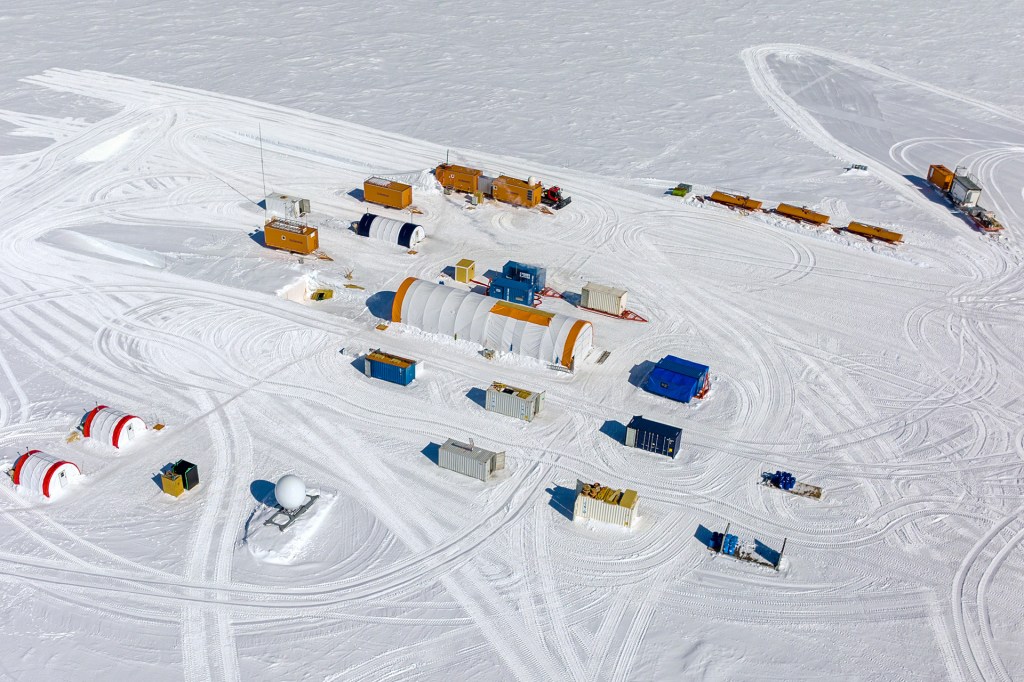 A small research camp with tents, containers, and equipment sits on a wide, flat sheet of snow.