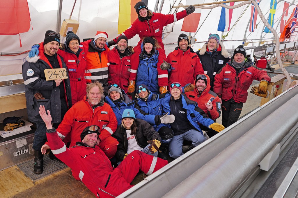 A large group of researchers in red and blue cold-weather clothing pose together inside a work shelter.